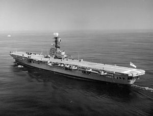 Side view of an aircraft carrier in motion. A helicopter sits on the carrier's deck, and several dark-uniformed people can also be seen.